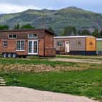 Des tiny houses en bois installées sur de vastes terrains verts, entourées d'arbres fruitiers, avec des montagnes en arrière-plan sous un ciel bleu clair