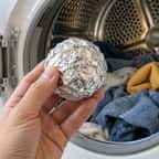 Une main tenant une boule de papier aluminium devant le hublot ouvert d'un sèche-linge rempli de vêtements