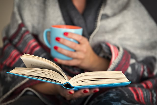 Une femme qui lit un livre, une tasse dans la main, un plaid autour des épaules