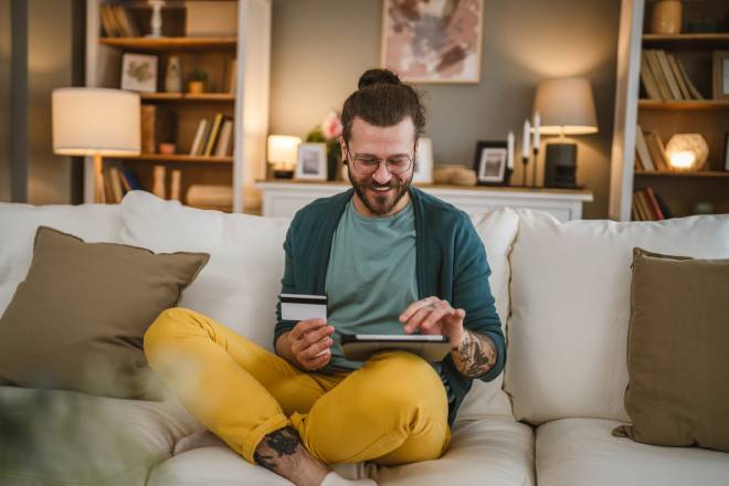 Homme tenant sa carte de cr&eacute;dit et faisant des achats depuis son canap&eacute;